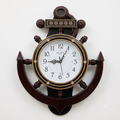 A decorative nautical wall clock shaped like an anchor, featuring a dark brown wood finish, gold-toned accents, and a white clock face with black numerals.